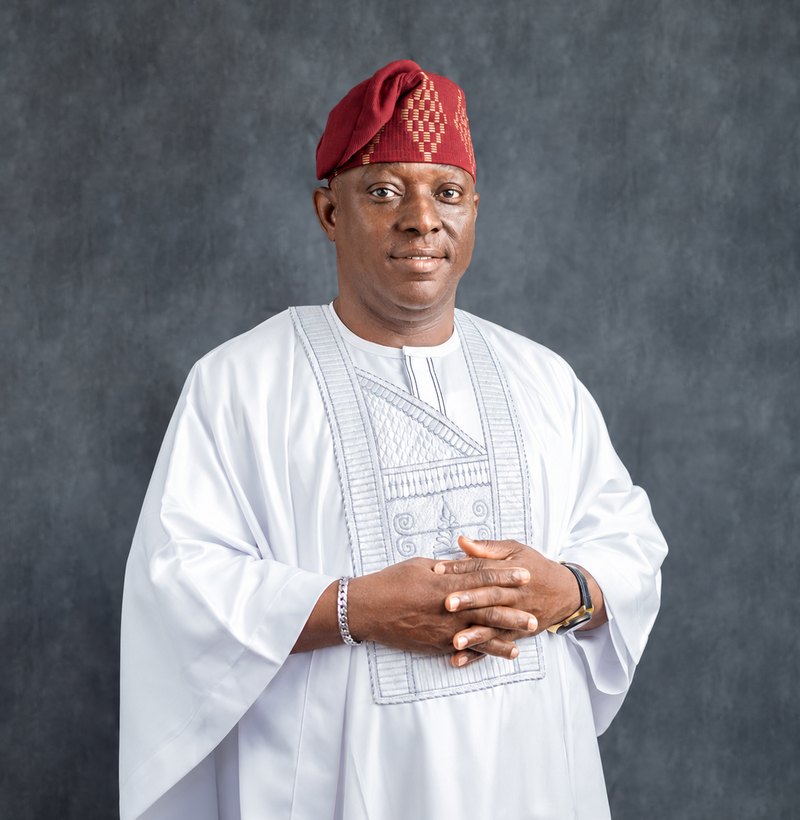 Kunle Olatunji in a white agbada and red cap, hands clasped, looking at the camera